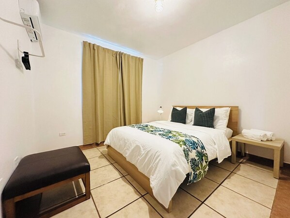 A hotel room with a bed, a bench, a nightstand, and a window with curtains.