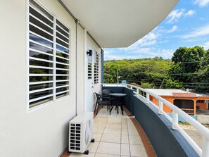Un balcon avec une table et des chaises, un climatiseur blanc et une vue sur un paysage verdoyant.