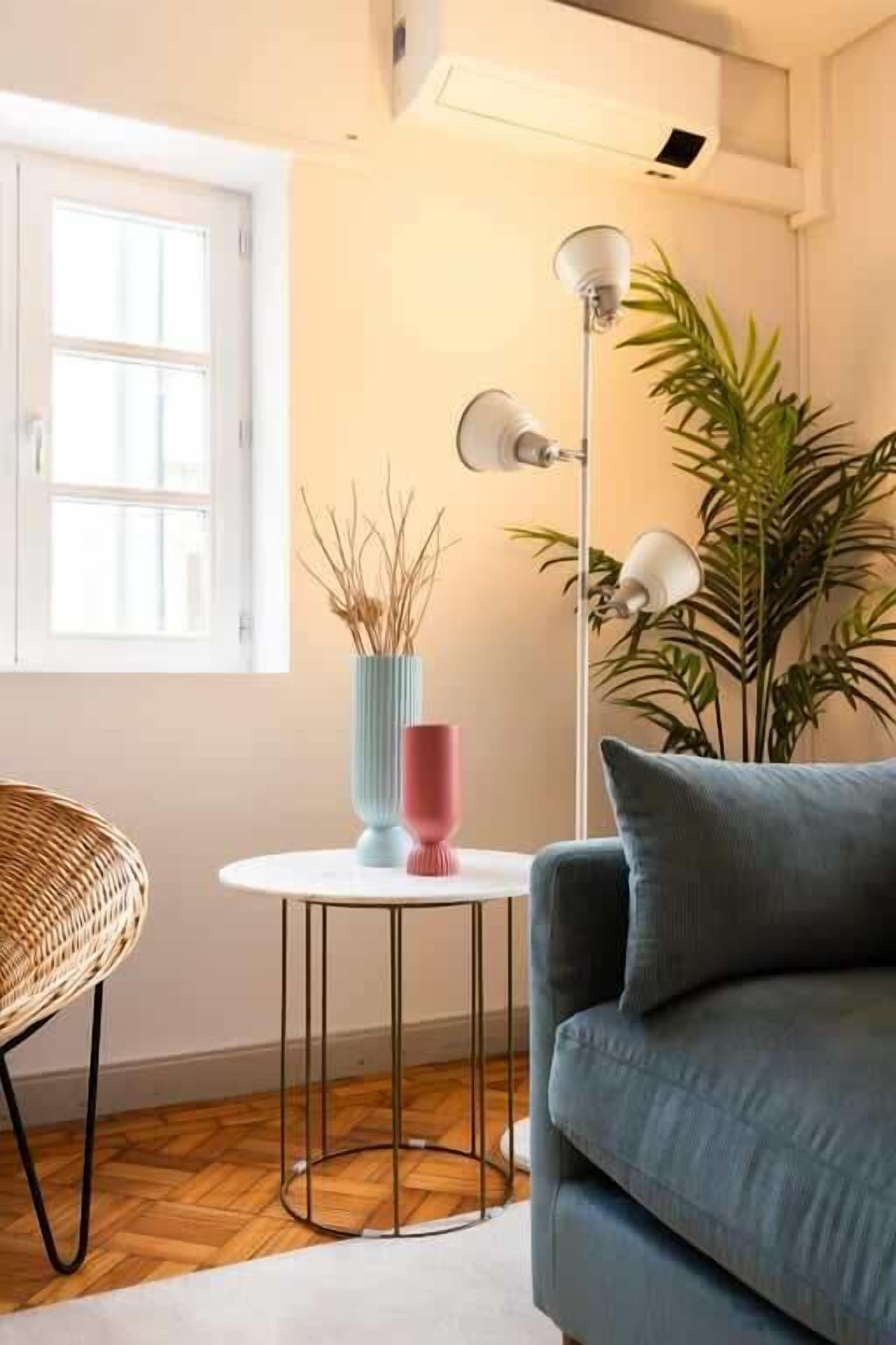Una habitación con un sofá azul, una mesita redonda con un jarrón y una lámpara, una silla de mimbre y una planta grande.