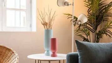 Una habitación con un sofá azul, una mesita redonda con un jarrón y una lámpara, una silla de mimbre y una planta grande.