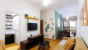 A living room with a sofa, a coffee table, a TV mounted on the wall, a bookshelf, and a dining area with a table and chairs.