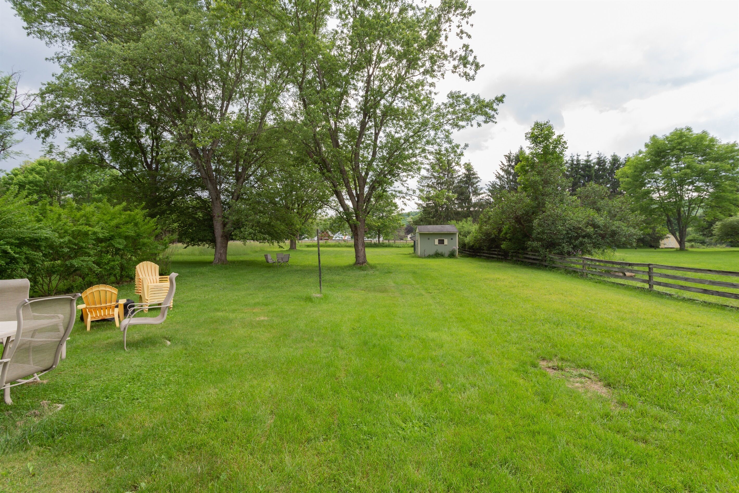 A grassy outdoor area with a small shed, a wooden fence, and Adirondack chairs.