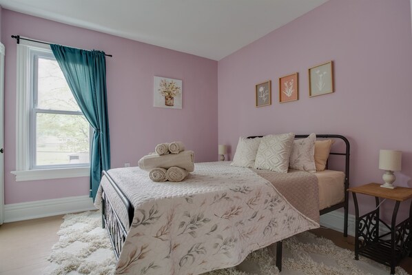 A bedroom with a bed, a window with curtains, a bedside table with a lamp, and wall art.