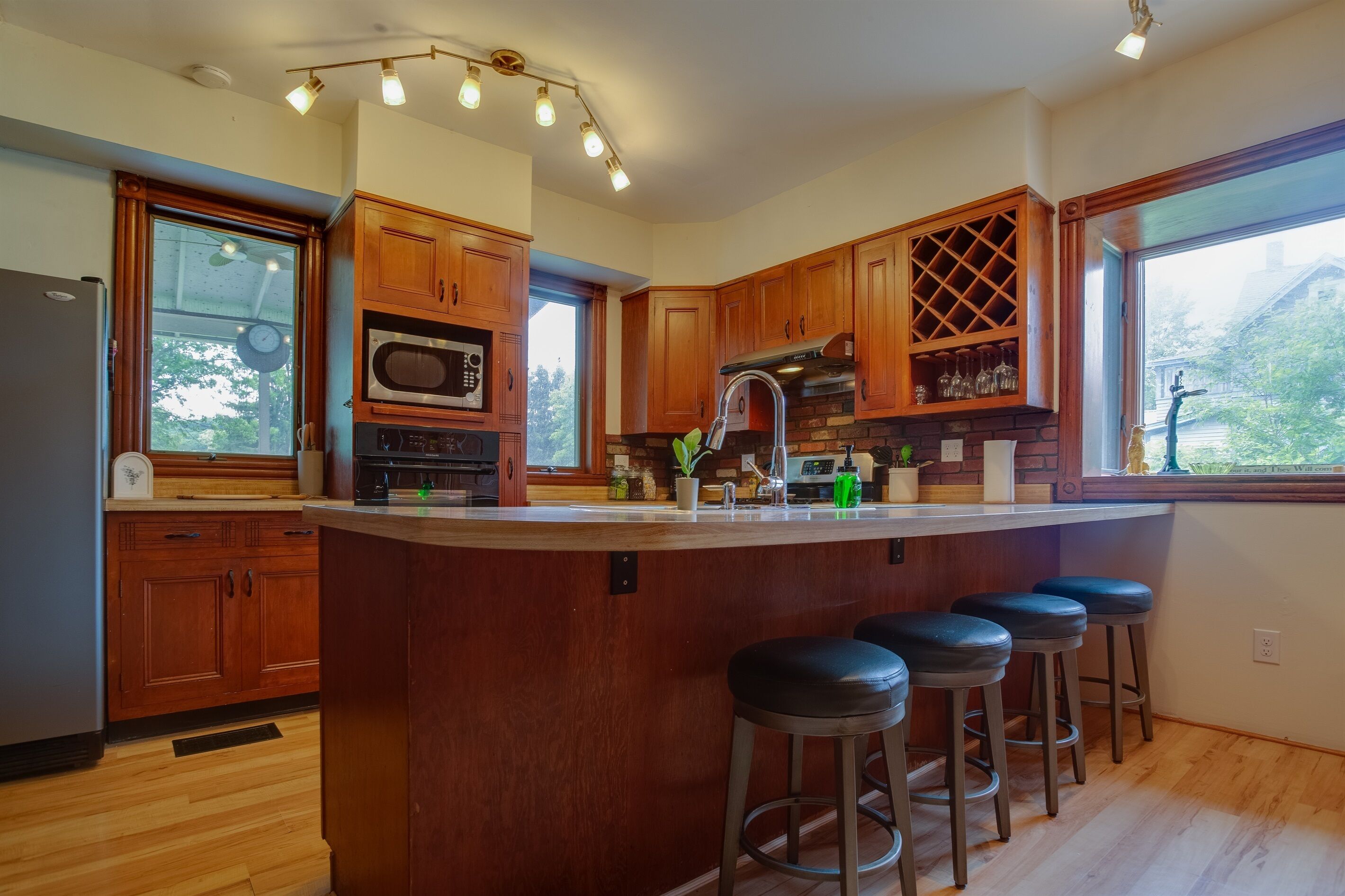 A modern kitchen with wooden cabinets, a central island with stools, a microwave, and a large window providing a view of the outdoors.