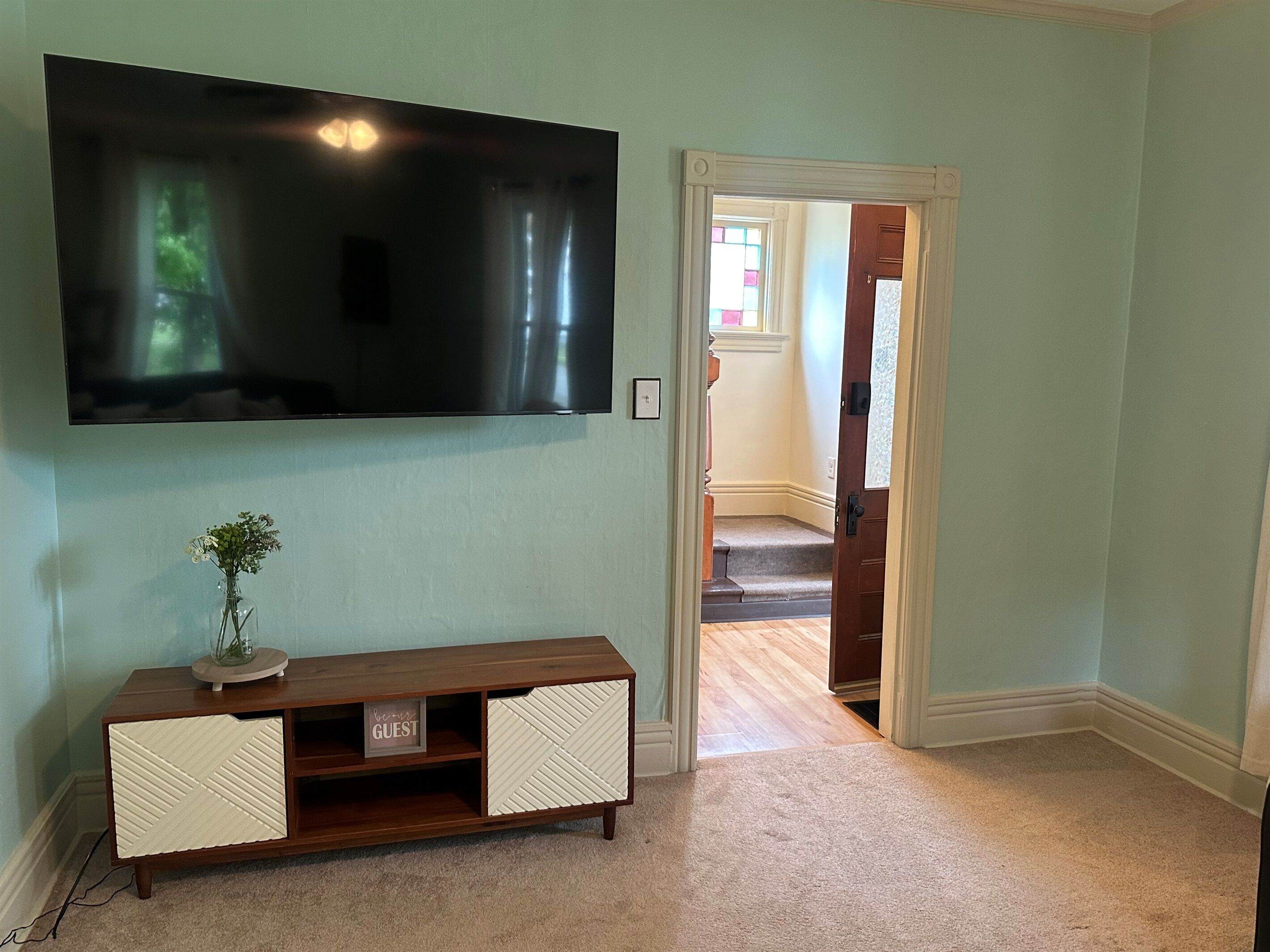 A room with a flat-screen TV mounted on the wall, a wooden console table with a vase of flowers, and a carpeted floor.