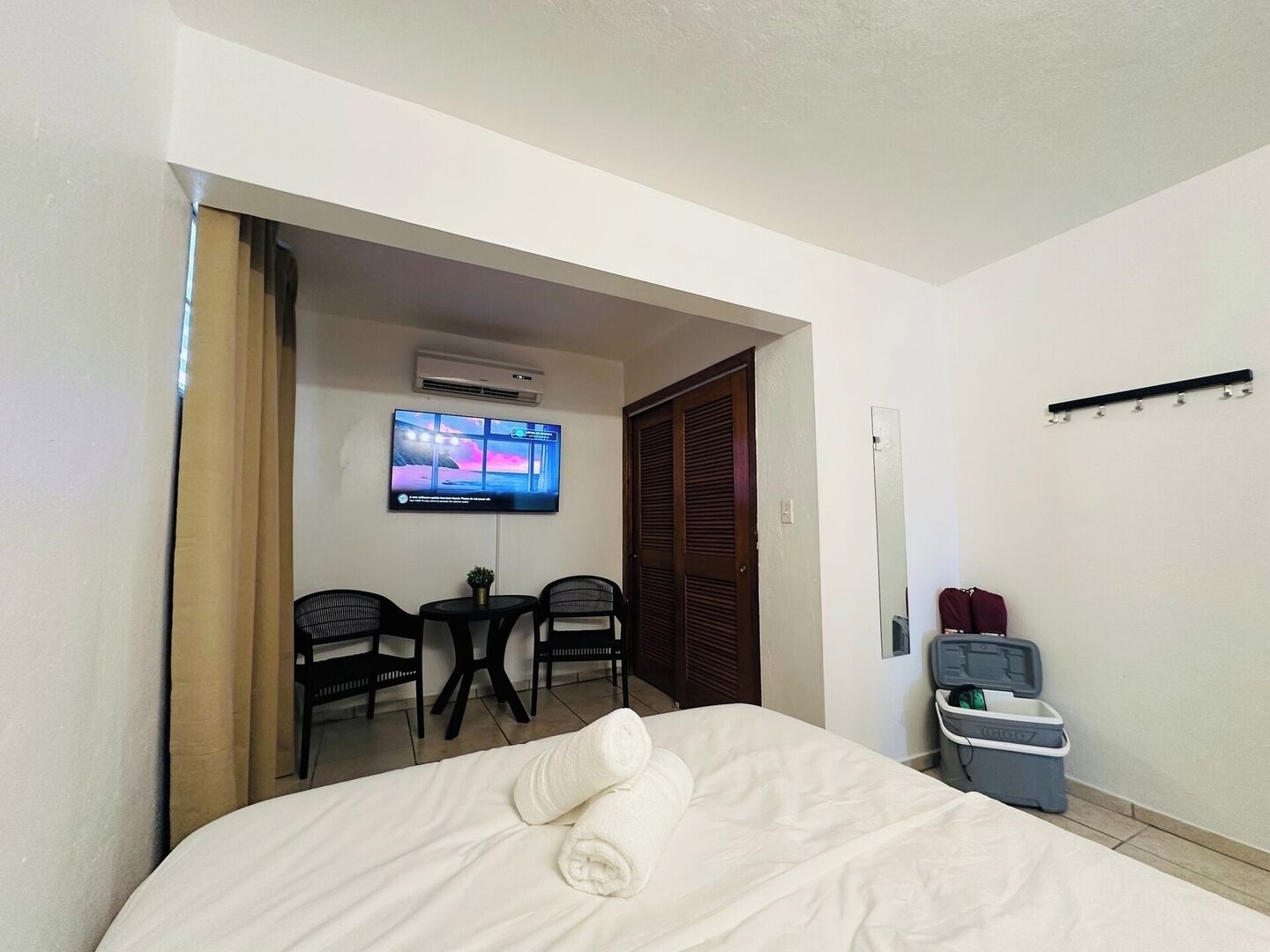 Habitación de hotel con cama, televisor montado en la pared, una mesita con dos sillas y un cesto de basura.