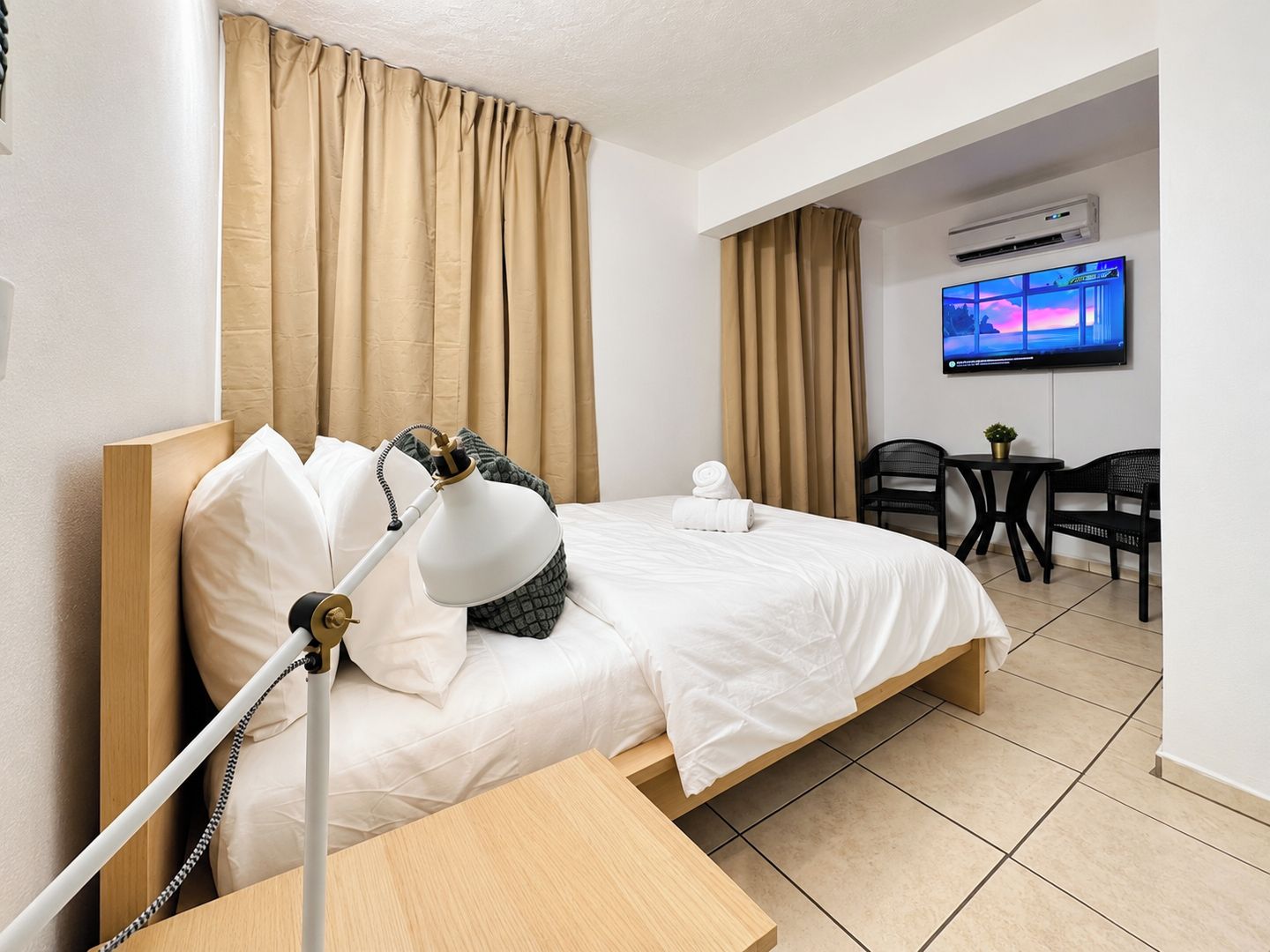 A hotel room with a bed, bedside lamp, curtains, a TV, a small table with a chair, and an air conditioning unit.