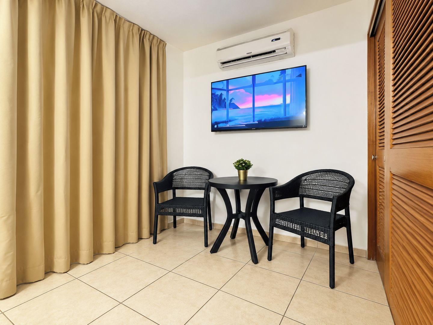 A room with a large window, a flat-screen TV, two chairs, a small table, and a potted plant.
