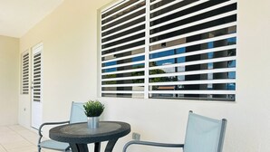A small outdoor seating area with a round table and two chairs, a potted plant, and a window with horizontal blinds. - Paradise Beach Apartment Vieques 3 (Vieques)