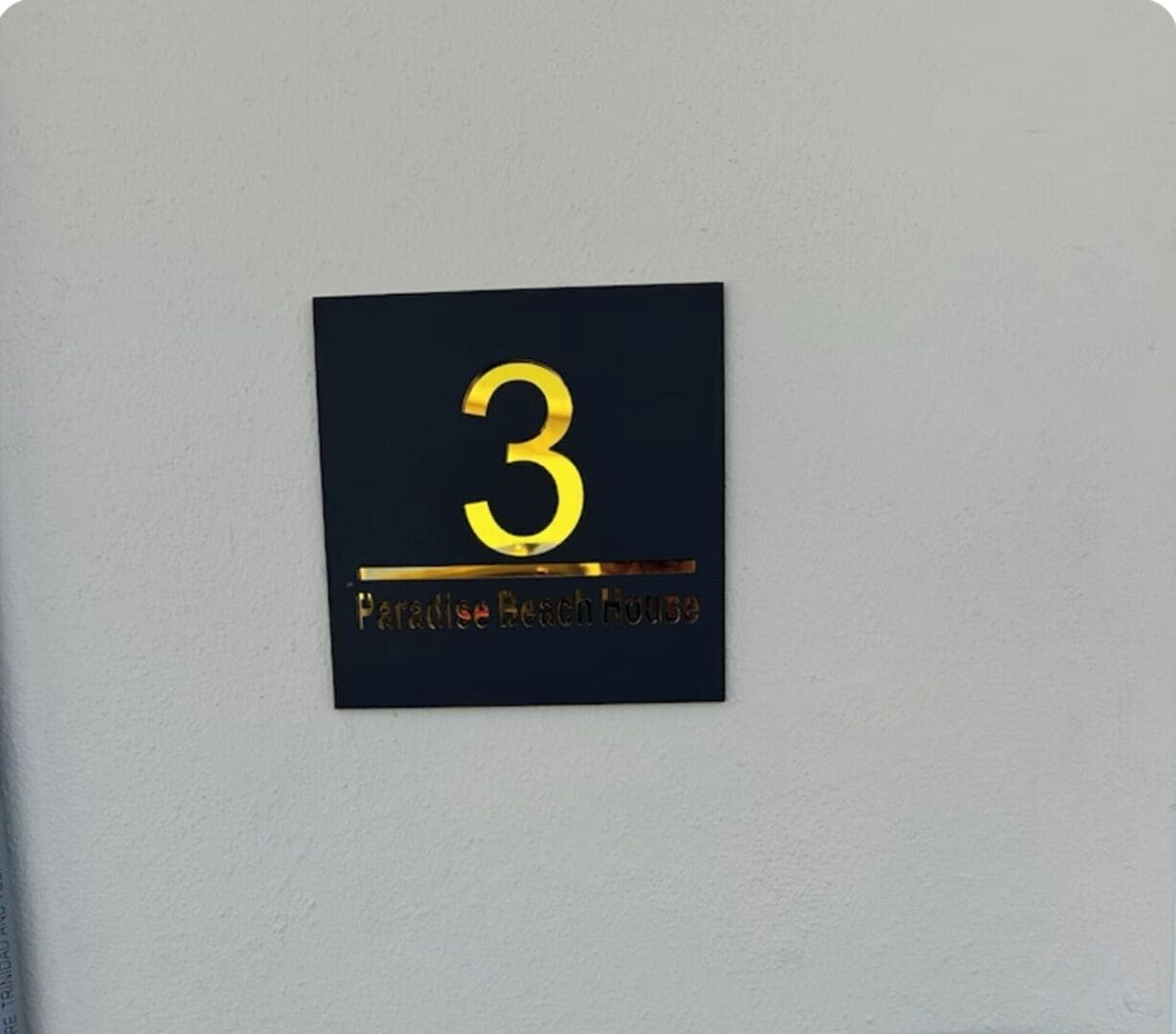 Ein schwarzes Schild mit der Nummer 3 und dem Schriftzug "Paradise Beach House" in Gold.
