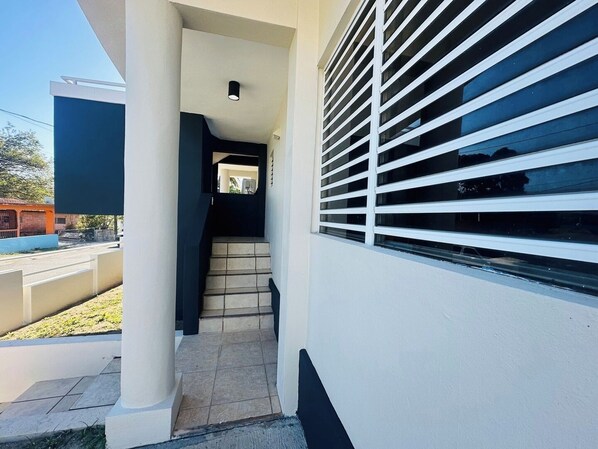 Un edificio moderno con una escalera que lleva a un nivel superior, un área exterior cubierta y una ventana con persianas horizontales.