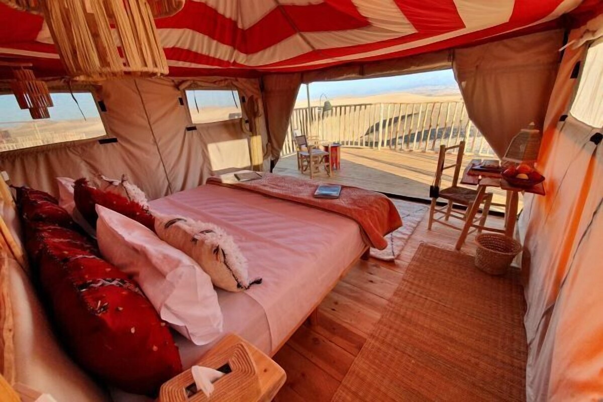 A tented room with a bed, pillows, a table with a chair, and a view of the desert.