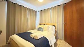 A neatly made bed with a blue and white duvet, two pillows, and rolled towels on the side. The room has beige curtains, a wooden door, and a ceiling light.