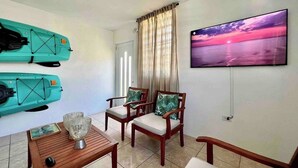A room with two kayaks, a wooden table with a glass, two chairs with cushions, a TV on the wall, and a window with curtains.