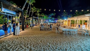 Un restaurant situé en bord de mer, avec une terrasse, des palmiers et des guirlandes lumineuses.
