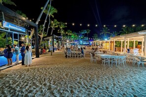 A beachside restaurant with outdoor seating, palm trees, and string lights.
