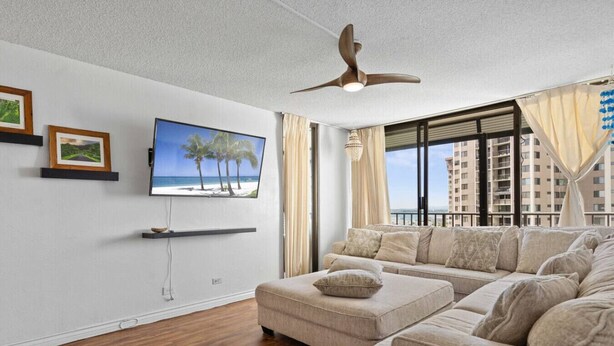 A living room with a large flat-screen TV, a ceiling fan, a sofa, and a window with a view of high-rise buildings.