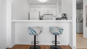 A compact kitchen with white cabinets, a white countertop, and a coffee machine. There are two bar stools with floral upholstery.