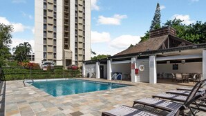 A swimming pool with lounge chairs, a covered patio, and a tall building in the background.