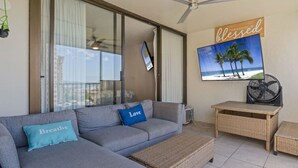 A modern living room with a grey sectional sofa, two cushions with text, a wicker side table, a ceiling fan, and a large TV screen displaying a beach scene.