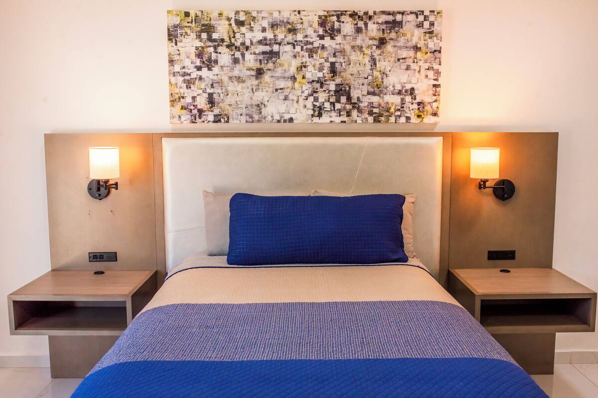 A hotel room with a large bed, two bedside tables, a headboard, and a wall-mounted abstract art piece.