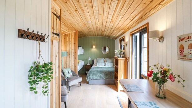 A cozy cabin interior with a wooden ceiling, a bed with a green bedspread, a dining table with a vase of flowers, and a wall-mounted lamp. - Woodpecker (Cambridge)