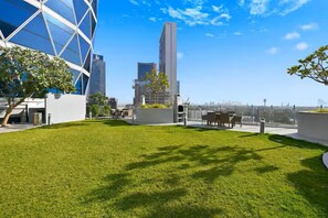A rooftop garden with a modern building, seating area, and a clear view of the city skyline.