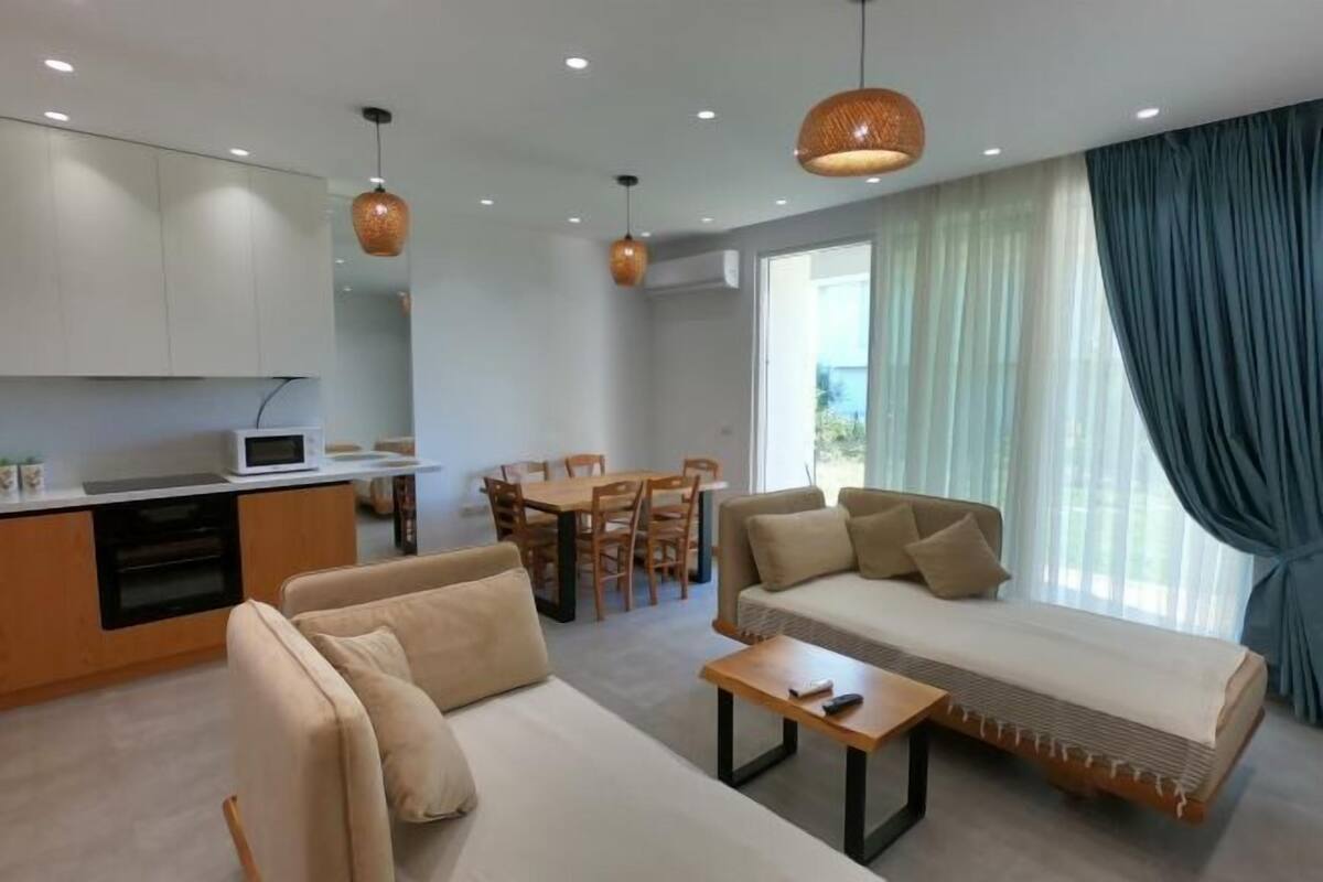 A modern living room with a sofa, a coffee table, a dining area with chairs, and a kitchen area with cabinets and a microwave.