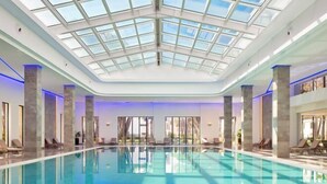 A large indoor swimming pool with a glass ceiling, surrounded by lounge chairs and a view of the outdoors. - Pinegrove Apartment (Durres)