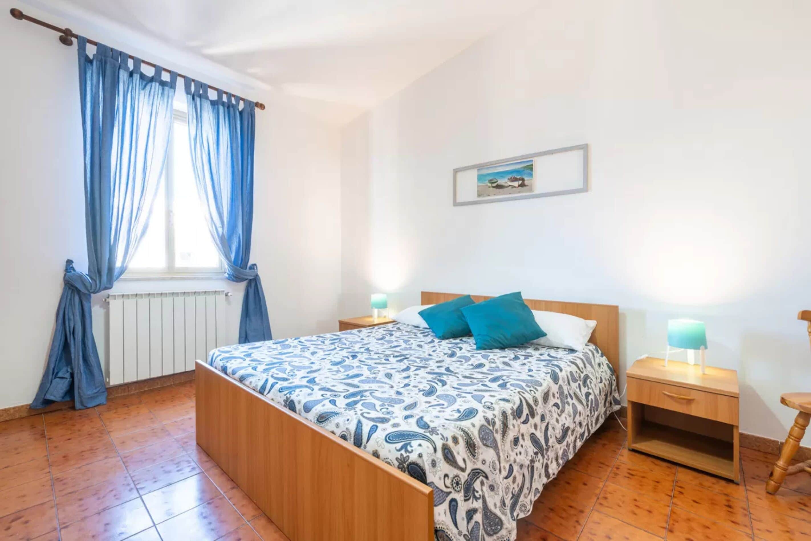 A bedroom with a wooden bed, blue curtains, a bedside table with a lamp, and a framed picture on the wall.