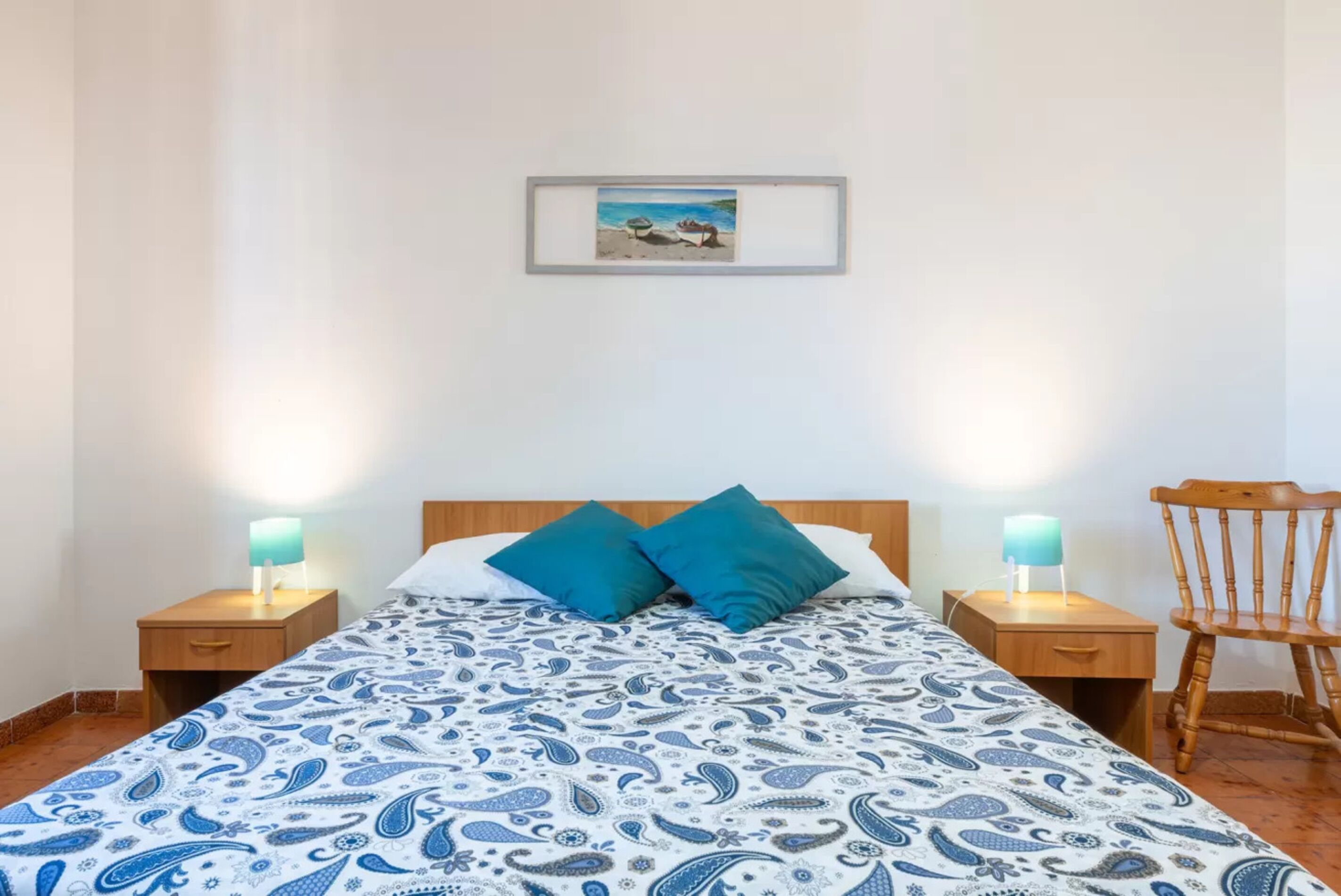 A neatly made bed with a patterned bedspread, two bedside lamps, a wooden nightstand, a wooden chair, and a framed picture on the wall.