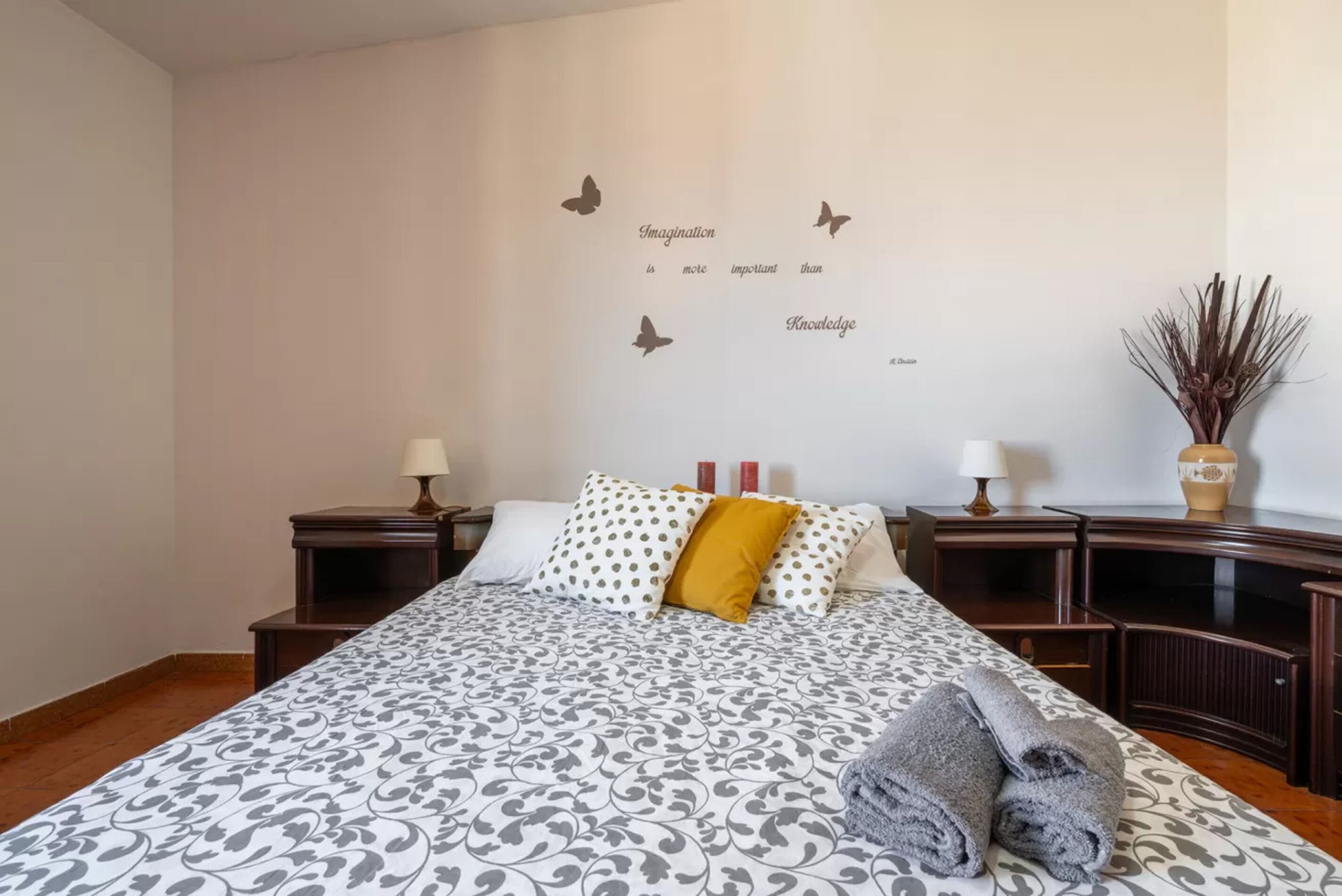 Una camera da letto ordinata con una coperta a motivi, una parete decorata con farfalle e una citazione, e un vaso con piante secche sul comodino.
