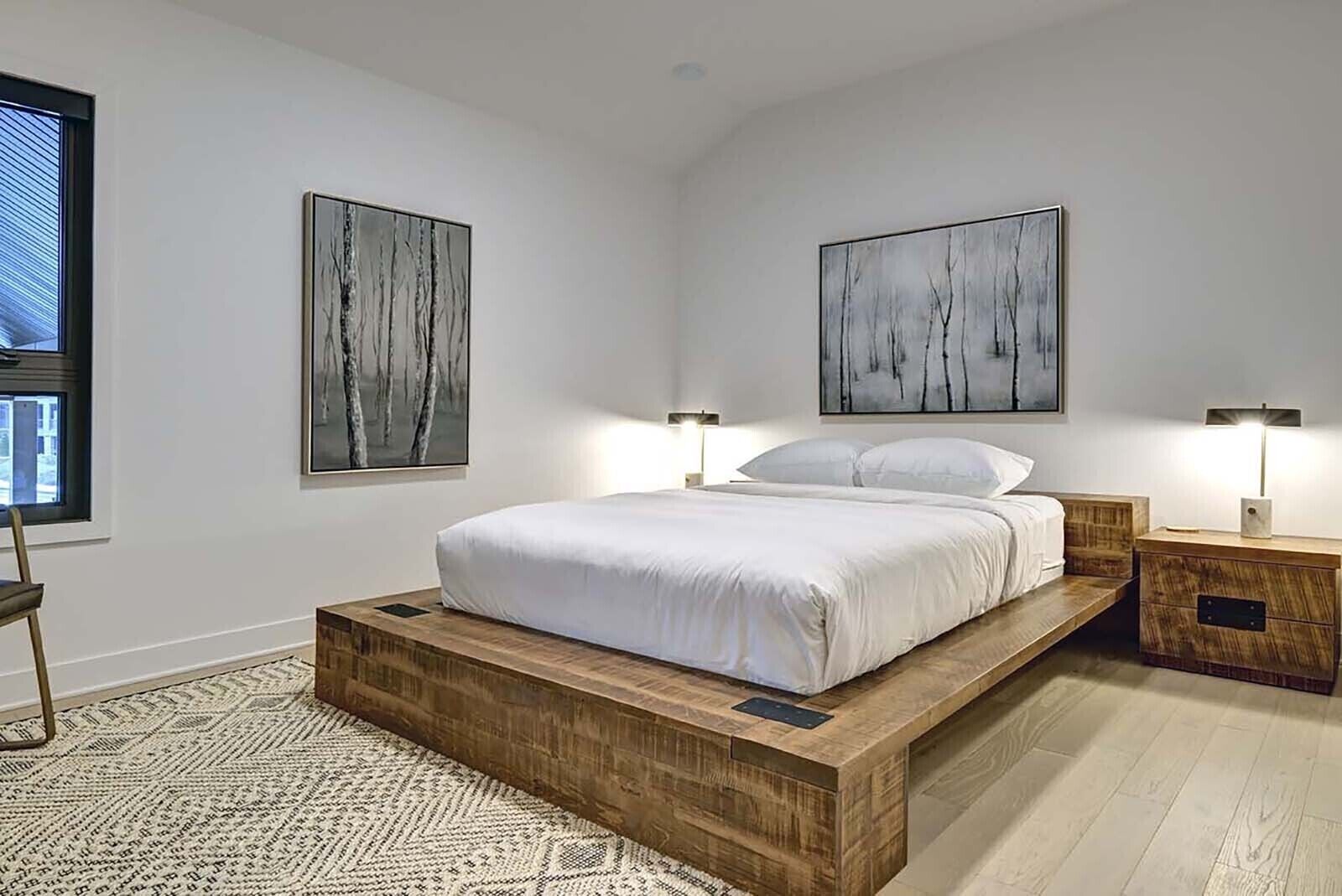 A modern bedroom with a wooden platform bed, two bedside tables, and two abstract paintings on the walls.