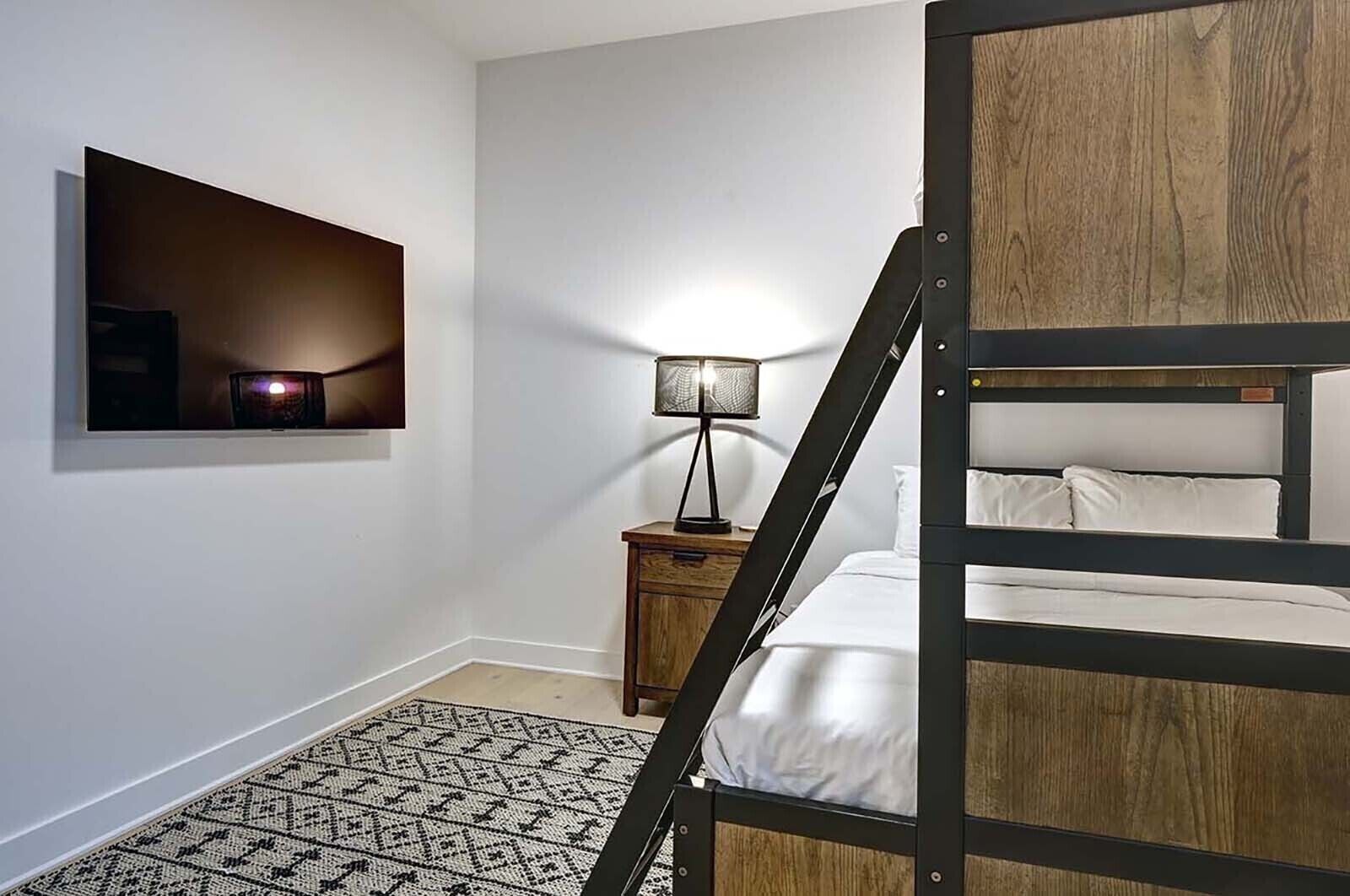 Un letto a castello con struttura in legno, un comodino con lampada e una mensola a muro con un quadro.