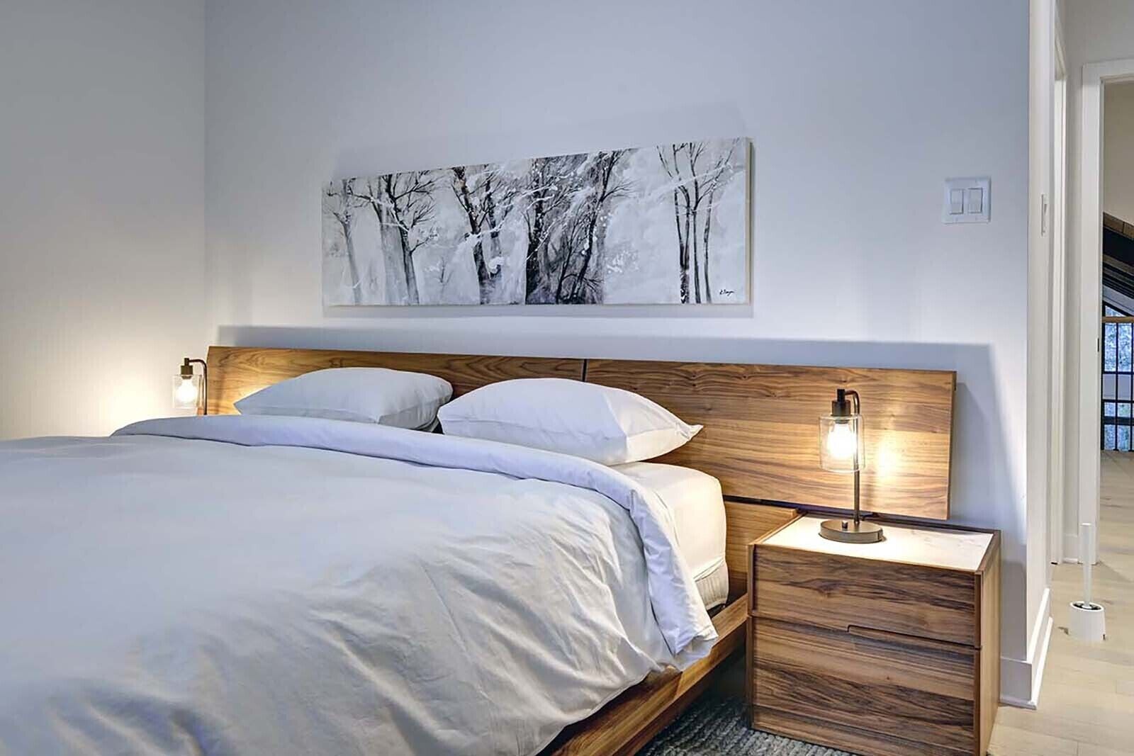 A bedroom with a wooden headboard, a bed with white bedding, a bedside table with a lamp, and a framed picture of trees on the wall.