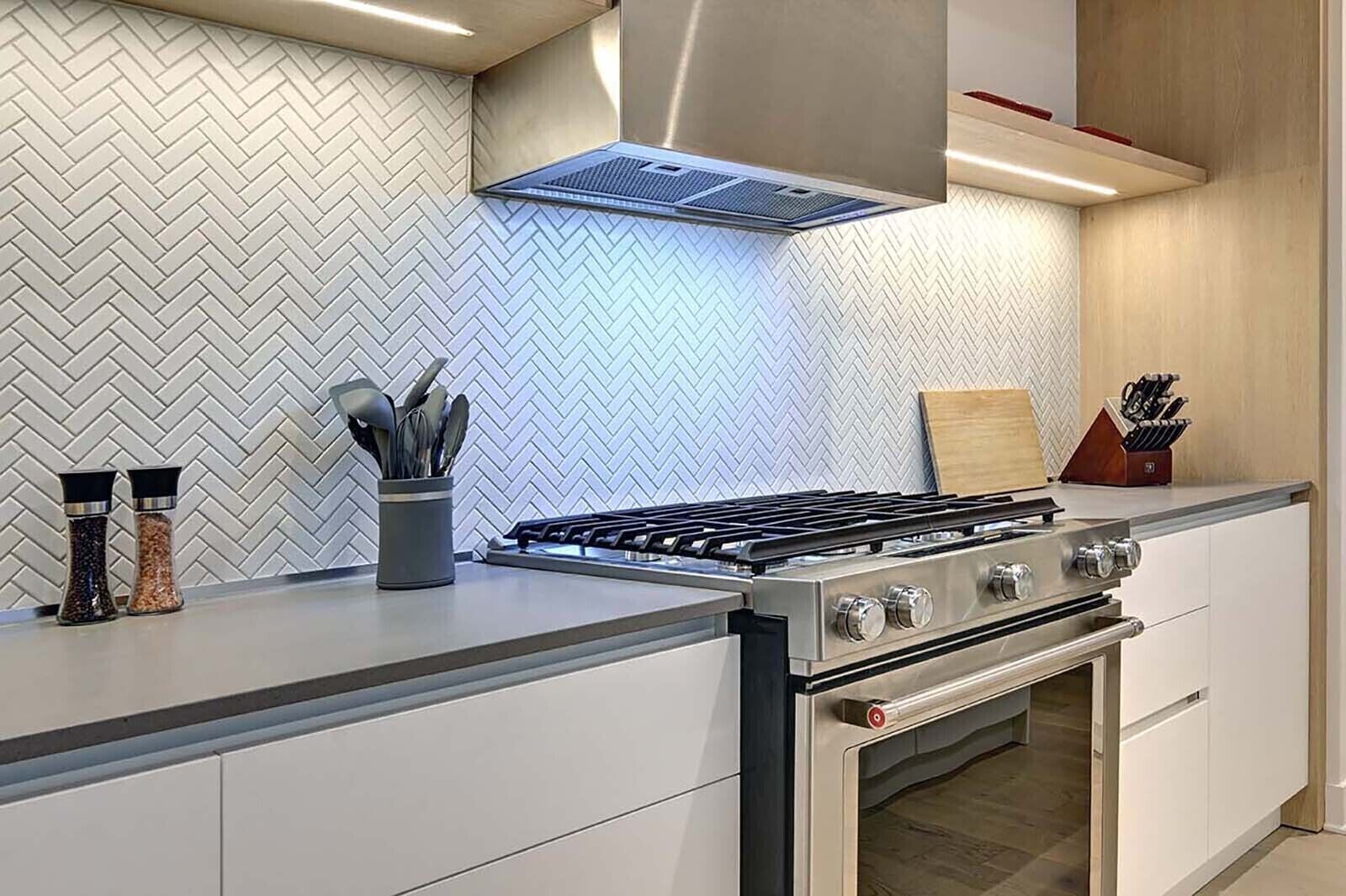 A modern kitchen with a stainless steel stove, white cabinetry, and a herringbone-patterned backsplash.