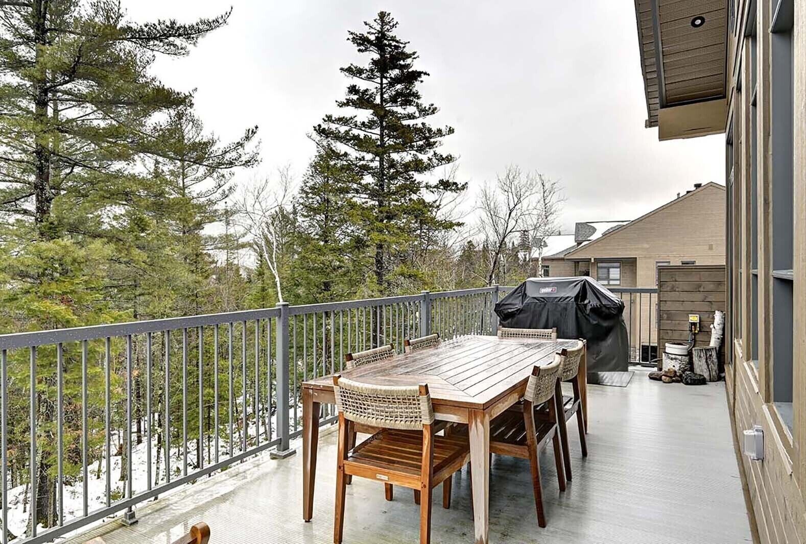Una terraza de madera con una mesa, sillas, una parrilla y vistas a árboles y un edificio.