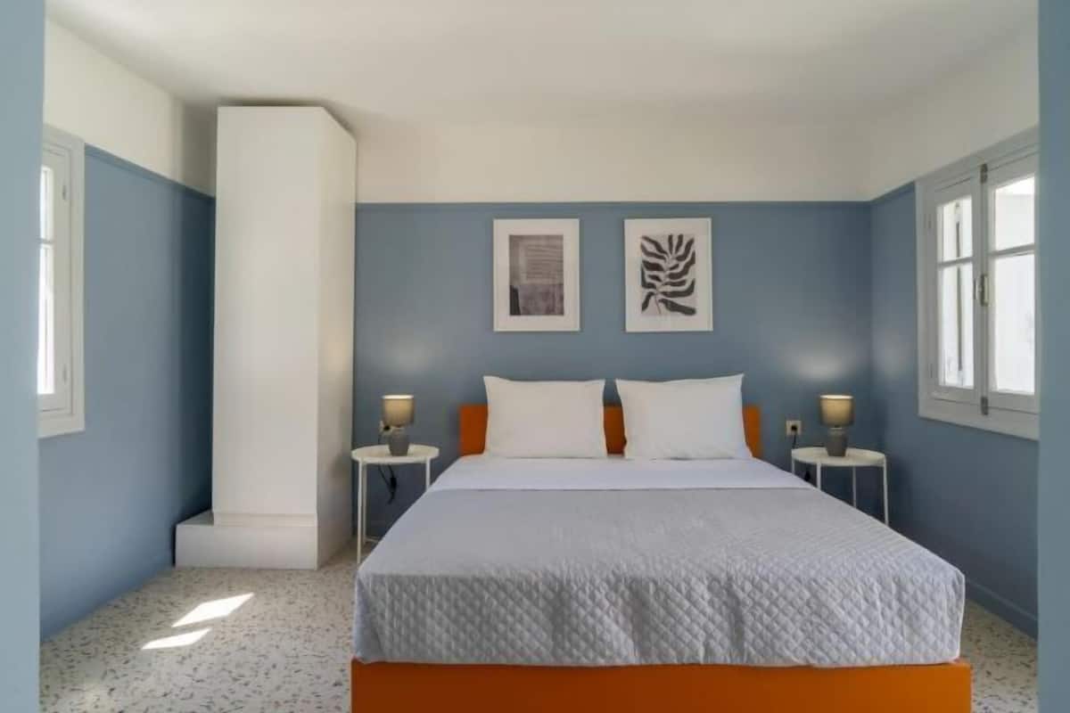 Une chambre à coucher avec un lit, deux tables de chevet, une fenêtre et des œuvres d’art accrochées au mur.