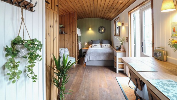A cozy room with a wooden ceiling, a bed with grey bedding, a bedside table with a lamp, and a window with a view. - The Heron (Cambridge)