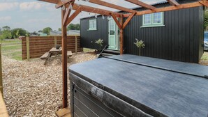 A wooden pergola with hanging light bulbs, a black shed with a green door, and a covered outdoor seating area. - The Heron (Cambridge)