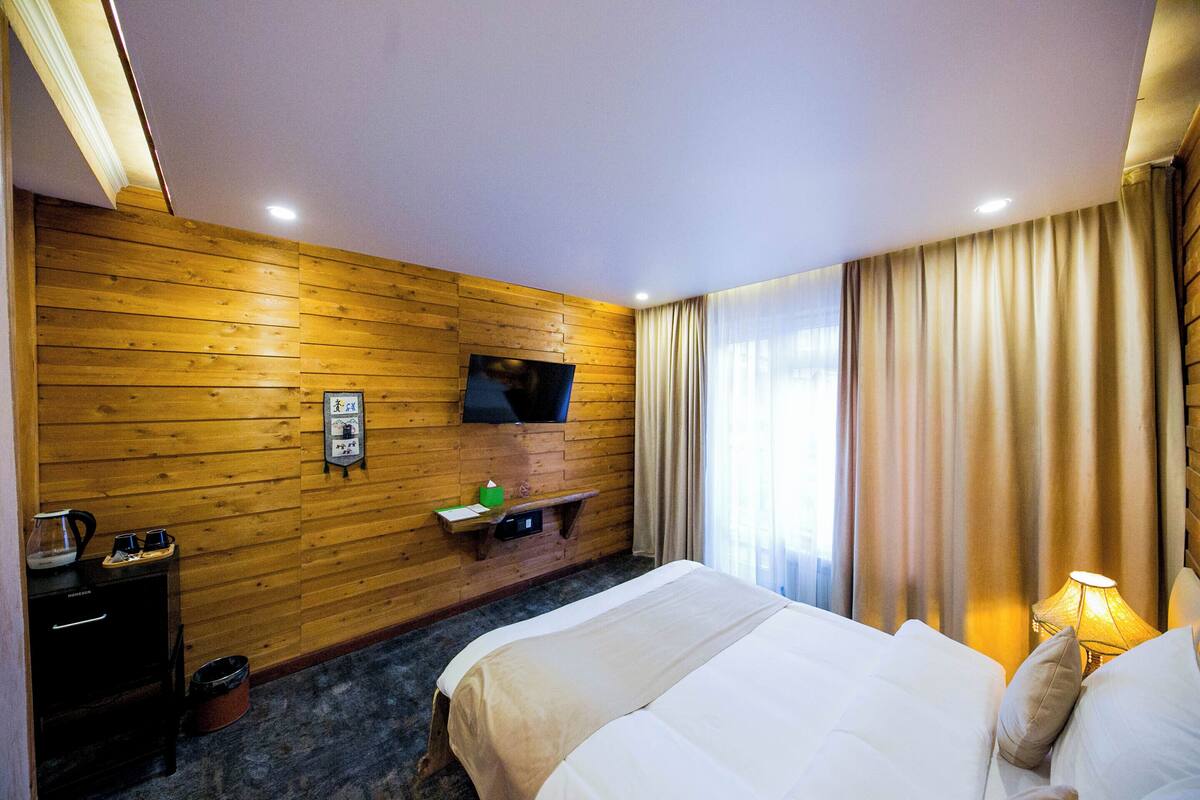 Una habitación de hotel con pared de madera, una cama, televisor montado en la pared, mesita de noche con lámpara y ventana con cortinas.