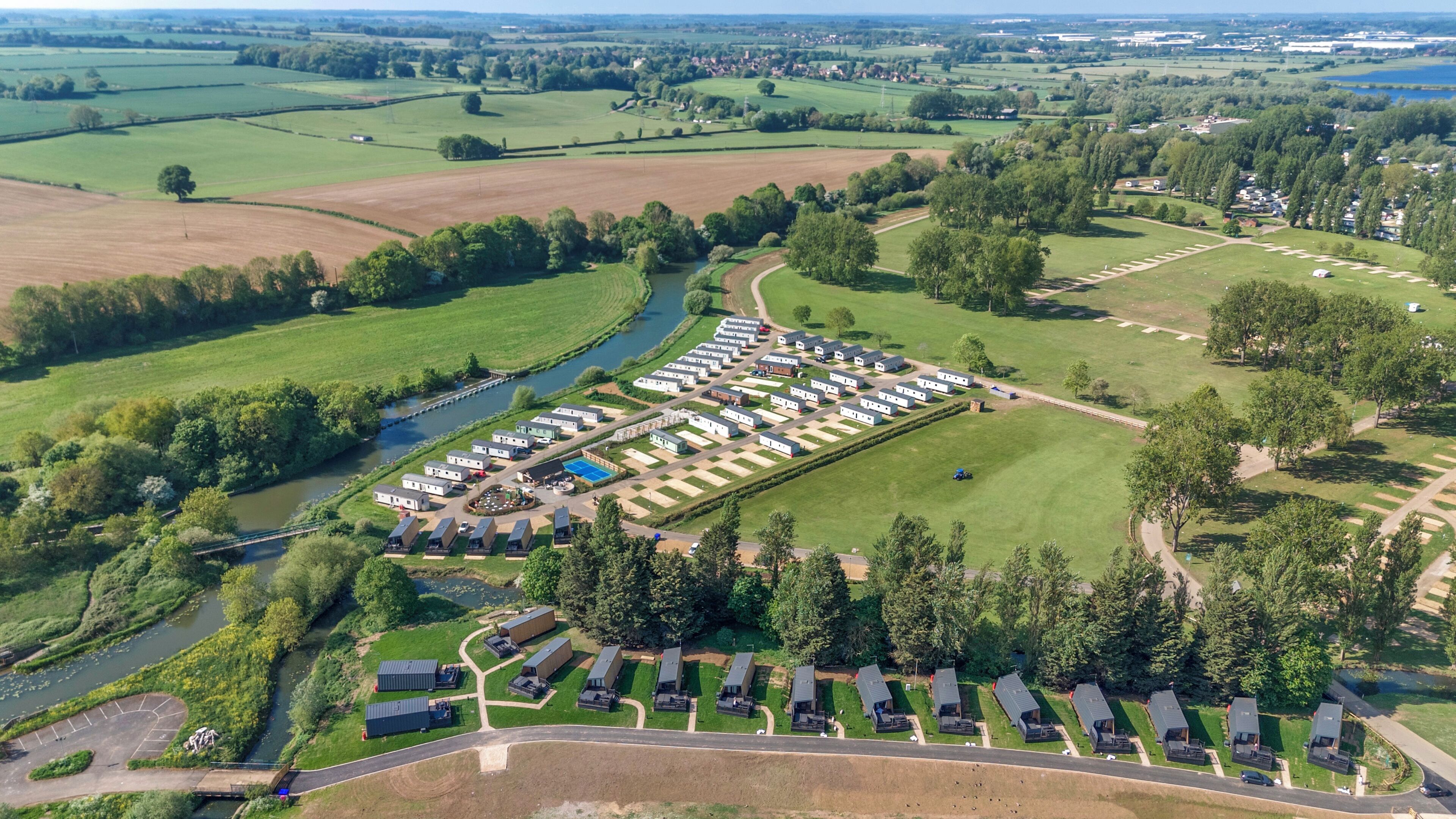 Vista aérea de un parque de caravanas con hileras de caravanas, un río y campos circundantes.