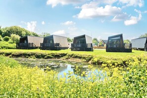 Une rangée de cabanes modernes et modulaires, dotées de terrasses en bois, est située au bord d’un plan d’eau paisible parsemé de nénuphars, et est entourée d’une végétation luxuriante.