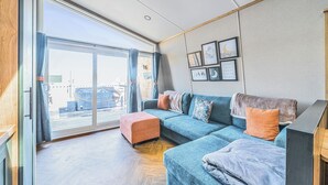Een moderne woonkamer met een blauwe bank, een oranje poef en een grote schuifglazen deur die leidt naar een buitenruimte.
