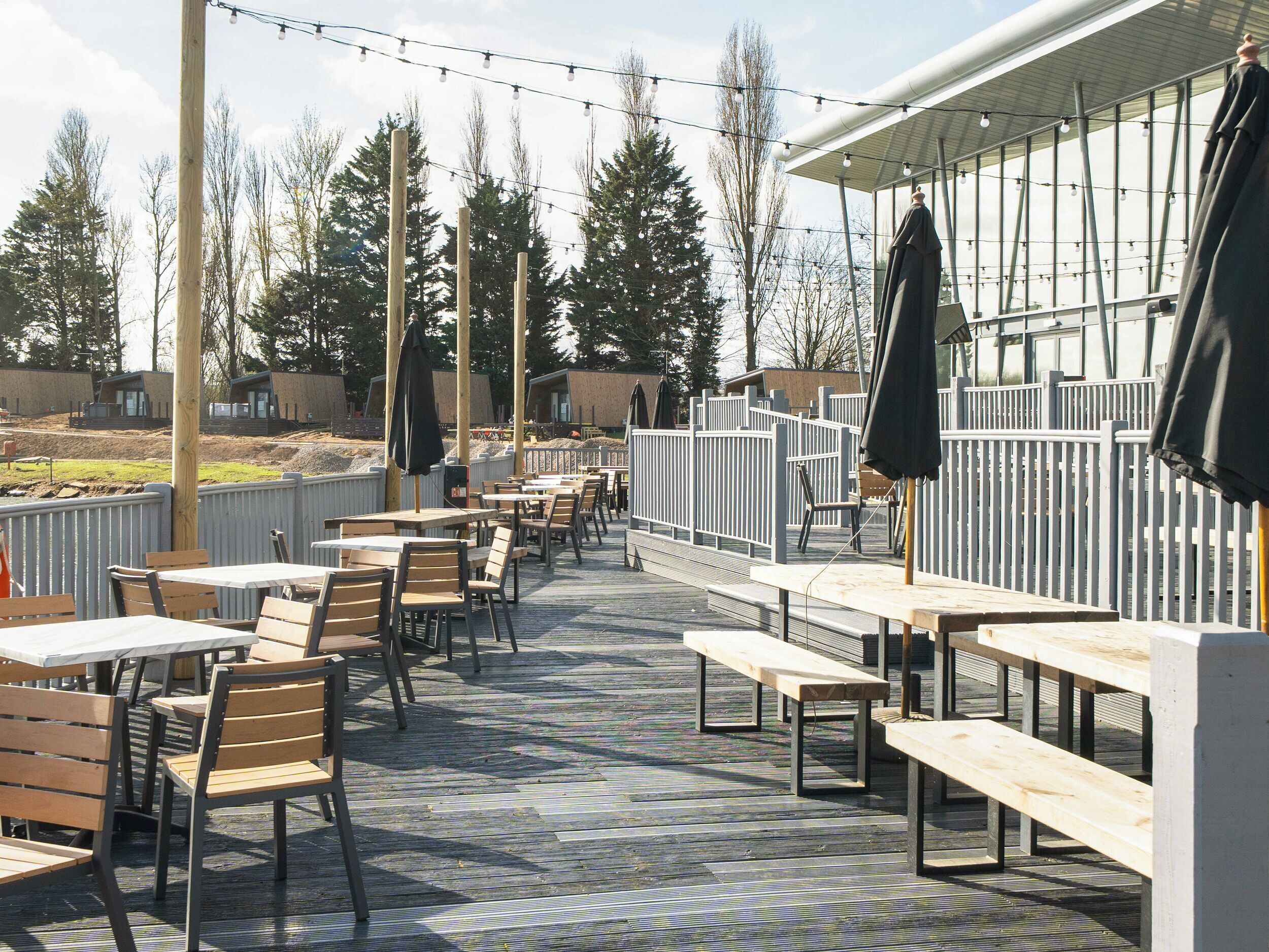 An outdoor patio with wooden tables and chairs, string lights, and a view of trees and buildings in the background.