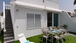 Ένα λευκό κτίριο με μπαλκόνι, μια τραπεζαρία με τραπέζι και καρέκλες και μια σκάλα που οδηγεί σε ανώτερο επίπεδο.