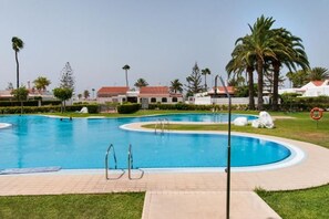A swimming pool with a clear blue water, surrounded by a paved deck, palm trees, and a building with a red roof in the background. - Bungalow JF/GI by VillaGranCanaria (Comarca Sur)