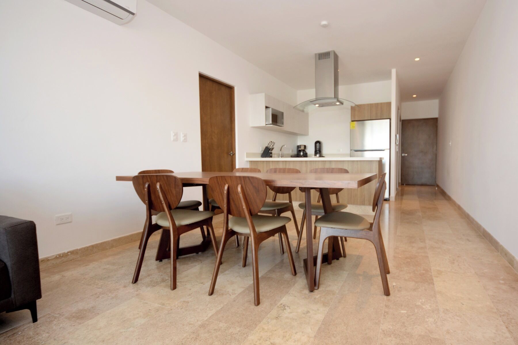 A modern kitchen with a central island, a dining area with six wooden chairs, and a small sofa in the foreground.