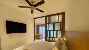 A hotel room with a bed, a ceiling fan, a flat-screen TV mounted on the wall, and a view of the dining area through a glass door. - Ipana Siempre Playa Dowtown (Playa del Carmen)