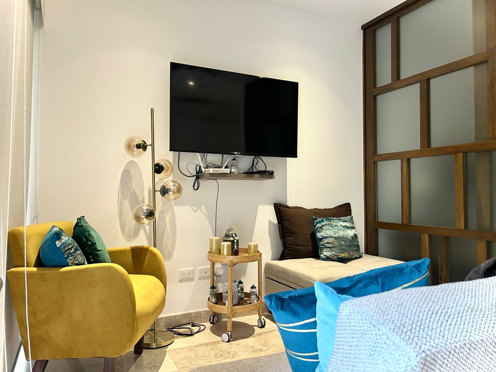 A modern living room with a yellow armchair, a sofa, a TV mounted on the wall, and a wooden-framed window.